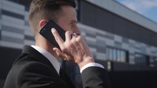 Atractivo hombre de negocios feliz hablando por teléfono móvil mientras está de pie cerca del edificio de oficinas . — Vídeos de Stock