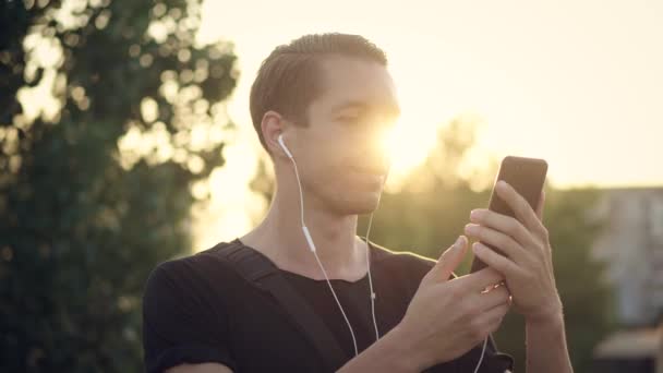 Genç adam şehirde günbatımında akıllı telefonunu kullanarak kulaklıkla müzik dinlemekten hoşlanır. — Stok video