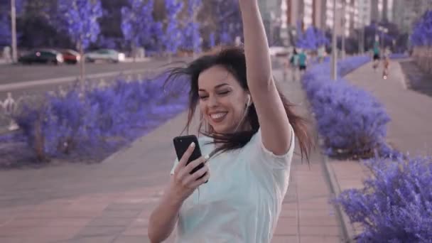 Mujer bastante joven disfrutando de su música favorita, bailando y caminando por la calle. Hermosa joven mujer sosteniendo el teléfono móvil escuchando música por auriculares en el fondo urbano de la ciudad . — Vídeos de Stock