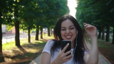 Neşeli, gülümseyen, kulaklıkla müzik dinleyen ve dans eden bir kadın. Tasasız genç kadın akıllı telefonu tutuyor ve gün batımında yürüyor, yavaş çekimde portreyi kapatıyor.