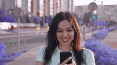 Elinde cep telefonu olan gülümseyen genç bir bayan, favori müziğinin tadını kulaklıkla çıkarıyor ve sokakta yürüyor. Sabit çekim.