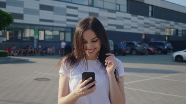 Portret trendy millennial jonge vrouw in hoofdtelefoon maakt gebruik van een smartphone op een stedelijke achtergrond. Glimlachende meisje student houdt smartphone in handen en geniet van de muziek in oortjes op de stadswandeling zonsondergang — Stockvideo