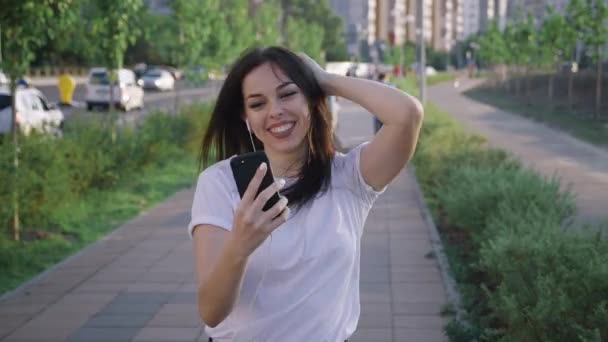 Mujer bastante joven disfrutando de su música favorita, bailando y caminando por la calle. Hermosa joven mujer sosteniendo el teléfono móvil escuchando música por auriculares en el fondo urbano de la ciudad . — Vídeo de stock