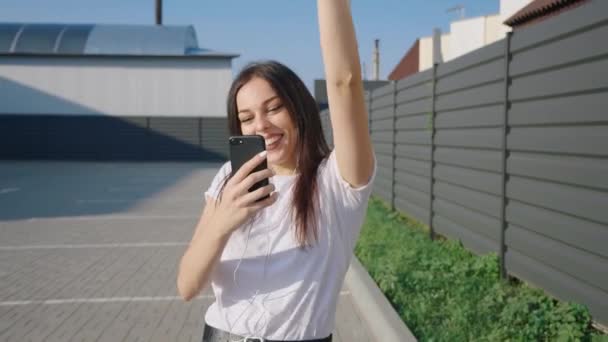 Usmívající se mladá žena drží mobilní telefon, raduje se z vítězství nebo vítězství a tance si užívá svou oblíbenou hudbu sluchátkem a kráčí po ulici. Záběr steadicamu. — Stock video