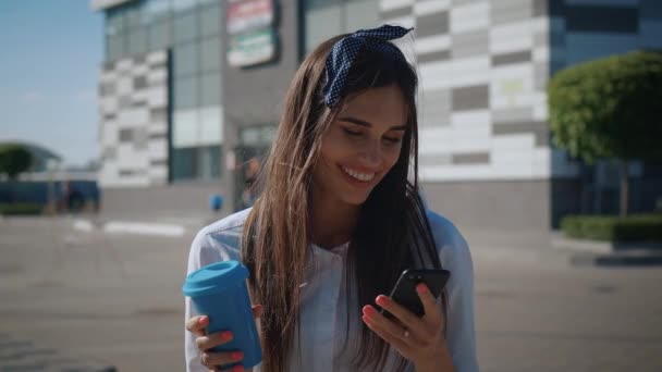 Улыбающаяся деловая женщина с помощью смартфона и кофе в центре города. Привлекательная молодая женщина наслаждается использованием телефона для социальных сетей на открытом воздухе . — стоковое видео