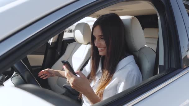 Vista lateral de la hermosa mujer sonriente sosteniendo el teléfono en el coche. Mujer de negocios sentada en un coche en el asiento del conductor y usando un teléfono inteligente — Vídeos de Stock