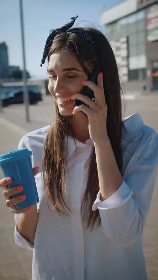 Verticale video van jonge vrouw praten aan de telefoon en het drinken van koffie op de achtergrond van kantoorgebouw. Schattig lachend meisje drinkt koffie en maakt gebruik van de telefoon op een stedelijke achtergrond. — Stockvideo