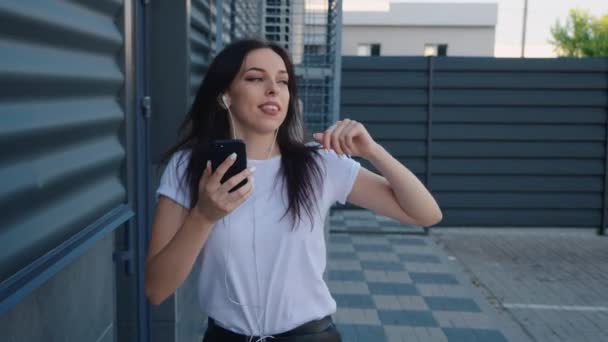 Retrato de una joven bailarina sonriente que escucha música con un smartphone sobre un fondo urbano. Chica bonita estudiante sostiene el teléfono inteligente en las manos y disfruta de la música con auriculares y bailando en una ciudad — Vídeos de Stock