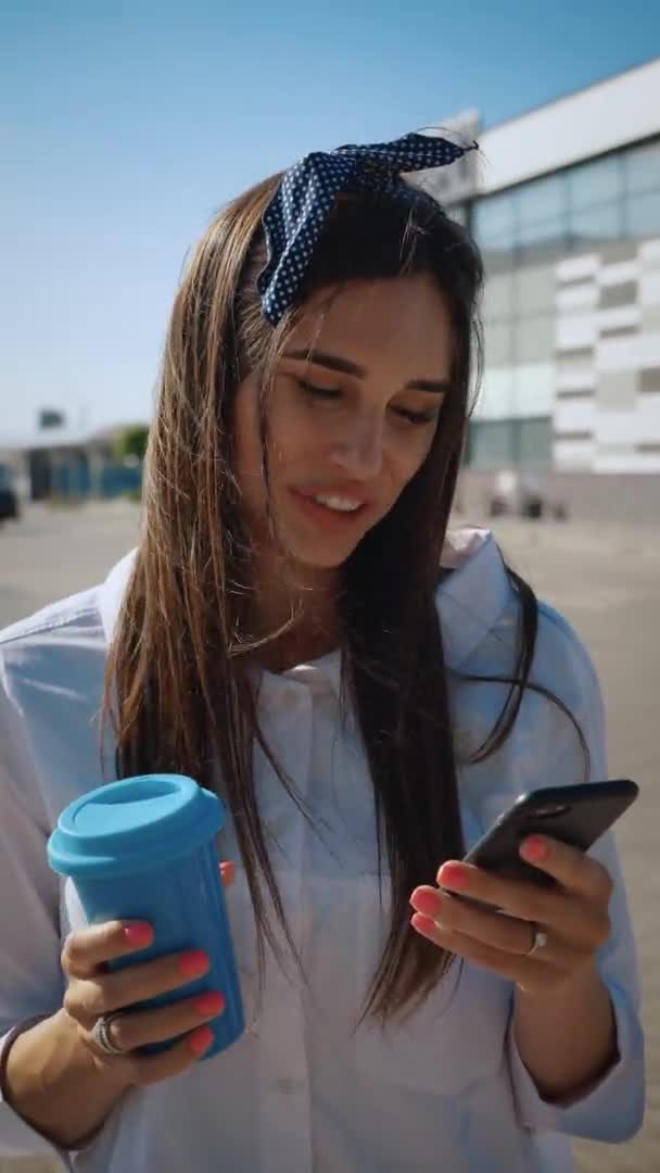 Vertikální video mladé ženy pomocí smartphonu a pití kávy na pozadí kancelářské budovy. Roztomilé usmívající se dívka pije kávu a používá telefon na městském pozadí. — Stock video