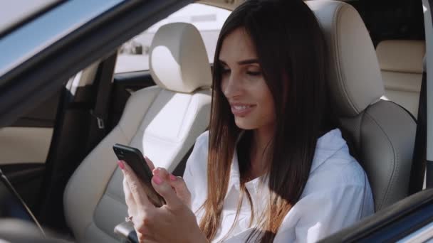 美しい若い女性が運転席に車の中で座って、スマートフォンを使用しています。笑顔のビジネス女性は車で携帯電話を保持. — ストック動画