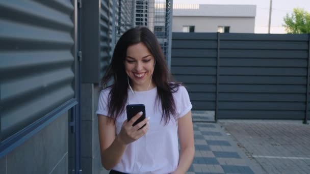 Portrét usměvavé mladé ženy se sluchátky pomocí smartphonu při chůzi po městské ulici. Šťastná dívka student drží chytrý telefon v rukou a má rád hudbu venku. — Stock video