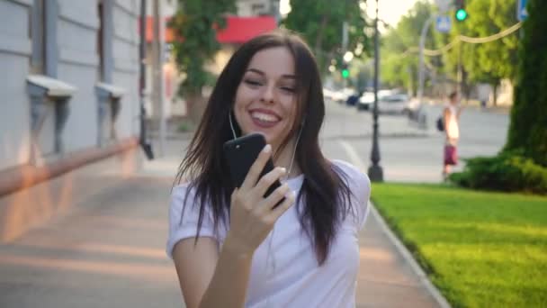Pretty young woman enjoying her favorite music, dancing and walking down the street. Beautiful young female holding mobile phone listening to music by earphone at city urban background. Steadicam shot — Stock Video