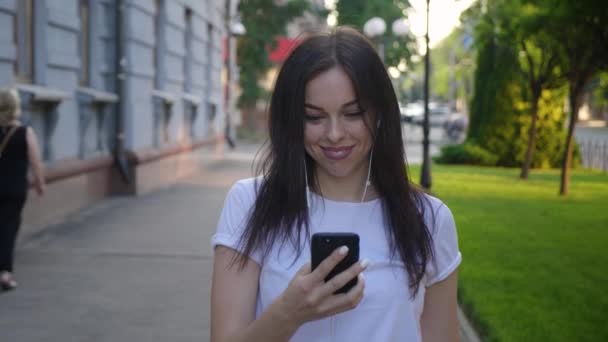 Portrait of smiling young woman with earphones using smartphone while walking on the city street. Happy girl student holds smart phone in hands and enjoys the music outdoors. — Stock Video