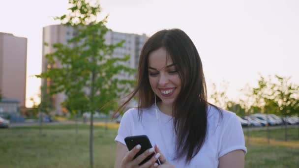 Портрет улыбающейся молодой женщины с помощью смартфона во время прогулки по городской улице на фоне заката. — стоковое видео