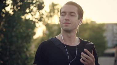 Genç adam şehirde günbatımında akıllı telefonunu kullanarak kulaklıkla müzik dinlemekten hoşlanır.
