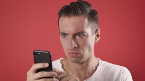 Angry Man Korzystanie ze smartfona na czerwonym tle. Człowiek jest zły na wiadomość, którą przeczytał w smartfonie. Angry man używa smartfona. — Wideo stockowe