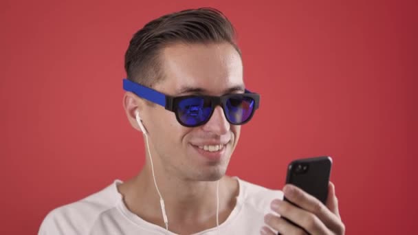 Fiatal hipster férfi napszemüveg élvezi hallgatni a zenét a fülhallgató segítségével okostelefon piros színű háttér — Stock videók