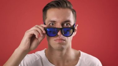 Şok olmuş ve kırmızı arka planda şaşırmış genç bir erkeğin portresi. Sürpriz yapan hippi adam güneş gözlüğü atıyor ve kameraya şaşkınlıkla bakıyor..