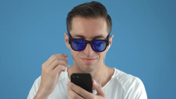 Joven hombre hipster en gafas de sol disfruta escuchando música en los auriculares utilizando su teléfono inteligente sobre fondo de color azul — Vídeo de stock