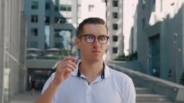 A megdöbbent fiatal üzletember portréja meglepetten veszi le a szemüvegét. Megdöbbent ember, akit megrémít a hír, a kamerába néz a város üzleti negyedének hátterében. — Stock videók