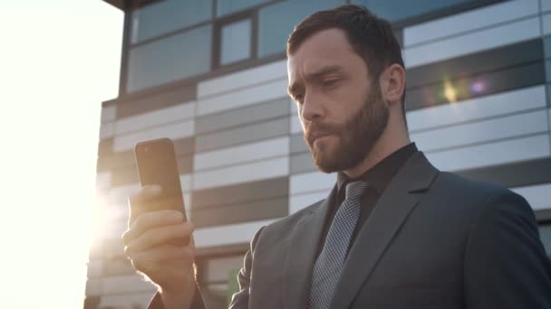 Portret van aantrekkelijke jonge zakenman met behulp van Smartphone staan in de buurt van Modern Office Building. Draagt een klassiek pak. Sociaal Netwerk. Apps. Smartphones. — Stockvideo