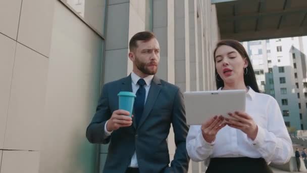 Un hombre de negocios y una mujer de negocios confiados están caminando por el centro y discutiendo negocios en el edificio de oficinas de fondo. Colegas hombre y mujer en el camino al trabajo están discutiendo el inicio con el uso de la tableta — Vídeo de stock