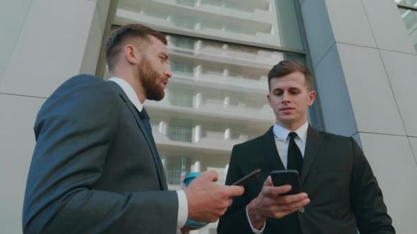 Два работника-мужчины взаимодействуют друг с другом в деловом районе города. Уверенные бизнесмены, использующие смартфон, обсуждают планирование бизнес-проекта в корпоративной командной работе — стоковое видео
