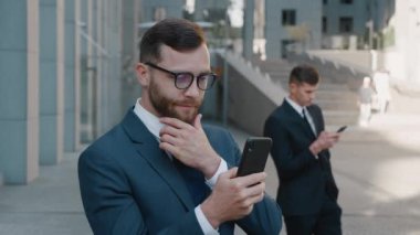 Klasik takım elbiseli şık sakallı iş adamının portresi modern ofis binasının yanında akıllı telefonunu kullanmaktan keyif alıyor. Gülümseyen beyaz erkek şehir merkezindeki iş bölgesinde mobil uygulama kullanıyor..