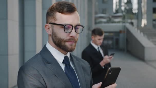 현대 사무실 건물 근처에서 스마트폰을 사용하여 정장을 하고 안경을 쓴 세련 된 수염을 가진 사업가의 초상화. 시내 사업 구역의 야외에서 모바일 앱을 사용하는 미소지어 지는 악어 수컷. — 비디오