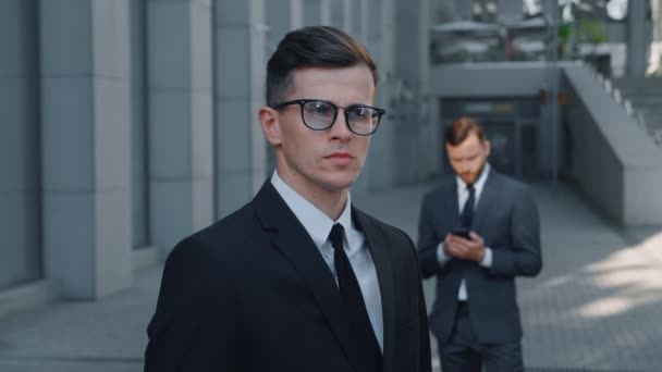 Egy vonzó üzletember arcképe öltönyben és szemüvegben, aki belenéz a belvárosi kamerába és a szabadban. Egy jóképű férfi üzletember portréja, aki az irodaház mellett áll — Stock videók