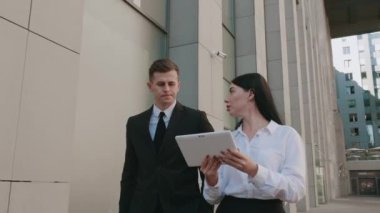 İş adamı ve iş kadını şehir merkezinde yürüyorlar ve ofis binasının arka planında iş konuşuyorlar. Erkek ve kadın meslektaşlar işe giderken tablet kullanımını tartışıyorlar.