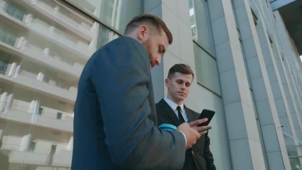 两名男性员工在商业区互相交流。有信心的商人使用智能手机讨论企业团队合作中计划的计算机业务项目头脑风暴 — 图库视频影像