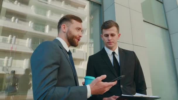 Two male employees interacting with each other in downtown business district. Confident businessmen using smartphone discussing planning computer business project brainstorm in corporate teamwork — Stock Video