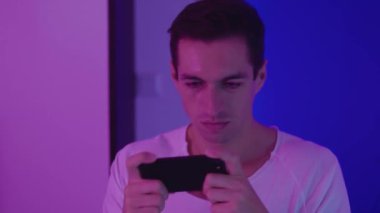 Akşamları neon ışıklarının üzerinde akıllı telefonuyla oyun oynayan bir adamın portresi. Genç erkek oyuncu mavi ve pembe neon ışıklı arka planda mobil uygulama kullanıyor