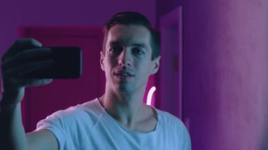 Akşamları neon ışıklarının üzerinde akıllı telefonuyla selfie çeken bir adamın portresi. Genç erkek sosyal medya için fotoğraf çekiyor mavi ve pembe neon ışıklı arka plan üzerinde mobil uygulama kullanıyor