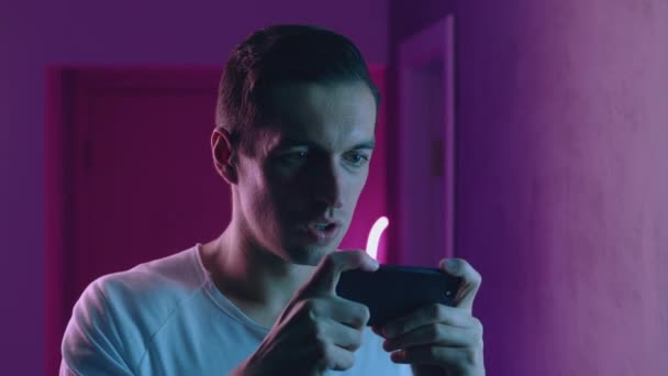 Retrato del hombre jugando juegos en su teléfono inteligente por la noche sobre luces de neón. Joven jugador masculino utilizando la aplicación móvil sobre fondo de luz de neón azul y rosa — Vídeo de stock