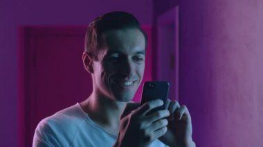 Neon ışıklarının üzerinde gece akıllı telefon kullanan gülümseyen bir adamın portresi. Genç beyaz erkek mavi ve pembe ışıklı arka planda mobil uygulama kullanıyor.