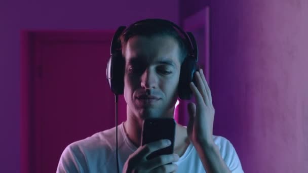 Portrait of smiling man enjoys listening to music with headphones in neon light. Young male listening to music using smartphone app on blue and pink neon light background — Stock Video