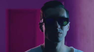 Gece kulübünün neon ışıkları altında hipster bir adamın portresi. Güneş gözlüklü genç erkek mavi ve pembe ışıklı arka planda kapalı bir yerde.