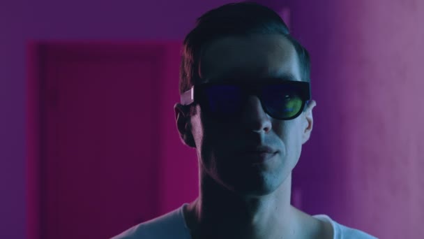 Gece kulübünün neon ışıkları altında hipster bir adamın portresi. Güneş gözlüklü genç erkek mavi ve pembe ışıklı arka planda kapalı bir yerde. — Stok video