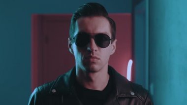 Gece kulübünün neon ışıkları altında vahşi bir adamın portresi. Ciddi erkek motorcu güneş gözlüklerini içeride mavi ve pembe ışıklı arka planda takıyor.