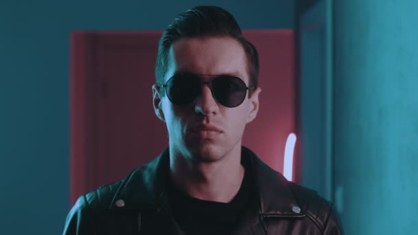 Portrait of brutal man inside at night over neon lights of nightclub. Serious male biker put on sunglasses indoors on blue and pink neon light background — Stock Video