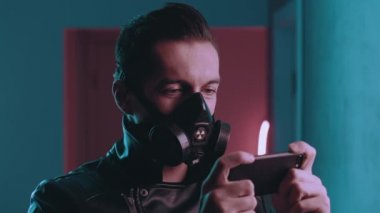 Solunum cihazındaki vahşi adamın portresi neon ışıkların üzerinde gece akıllı telefonuyla oyun oynuyor. Siber punk konsepti. Mavi ve pembe ışıklı arka planda mobil uygulama kullanan koruyucu maskeli bir erkek oyuncu