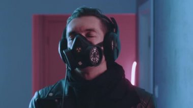 Solunum cihazındaki vahşi adamın siber punk portresi neon ışıkta kulaklıkla müzik dinlemeyi seviyor. Gaz maskeli genç erkek mavi ve pembe neon ışıkta müzik dinliyor..