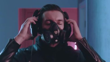 Solunum cihazındaki vahşi adamın siber punk portresi neon ışıkta kulaklıkla müzik dinlemeyi seviyor. Gaz maskeli genç erkek mavi ve pembe neon ışıkta müzik dinliyor..