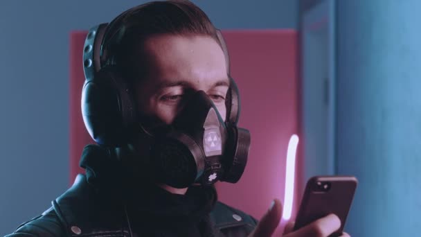 Cyberpunk portret van brutale man in respirator geniet van het luisteren naar muziek met koptelefoon op zijn smartphone in neon licht. man in gas masker luisteren naar muziek op blauw en roze neon licht achtergrond. — Stockvideo