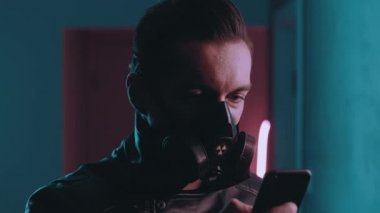 Geceleri neon ışıklarının üzerinde akıllı telefon kullanan tehlikeli bir adamın portresi. Siber punk konsepti. Mavi ve pembe ışıklı arka planda mobil uygulama kullanan koruyucu maskeli vahşi bir erkek.