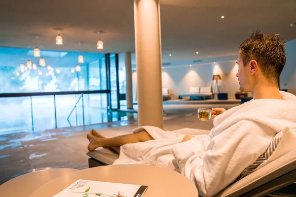 Saalbach hotel. Austria. March 20, 2018. Young man having a rest in the SPA by the open pool in a robe. Reading magazine.