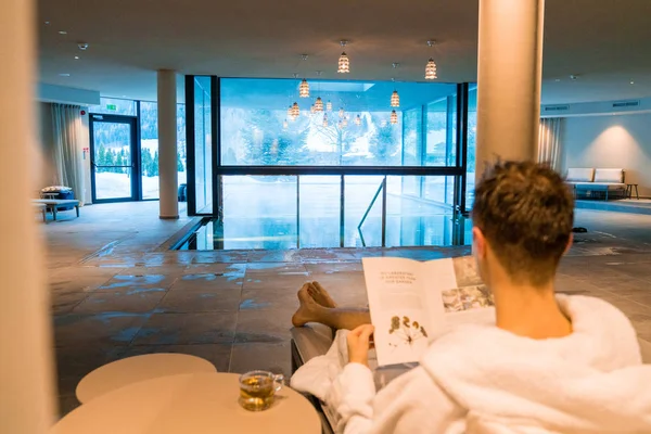 Saalbach hotel. Austria. March 20, 2018. Young man having a rest in the SPA by the open pool in a robe. Reading magazine.