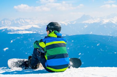 Saalbach, Avusturya. 20 Mart 2018. Genç adamla ufuk içine izlerken Alpleri'nde dağın tepesinde oturan bir snowboard. Kayak Merkezi hayran. Snowboard.